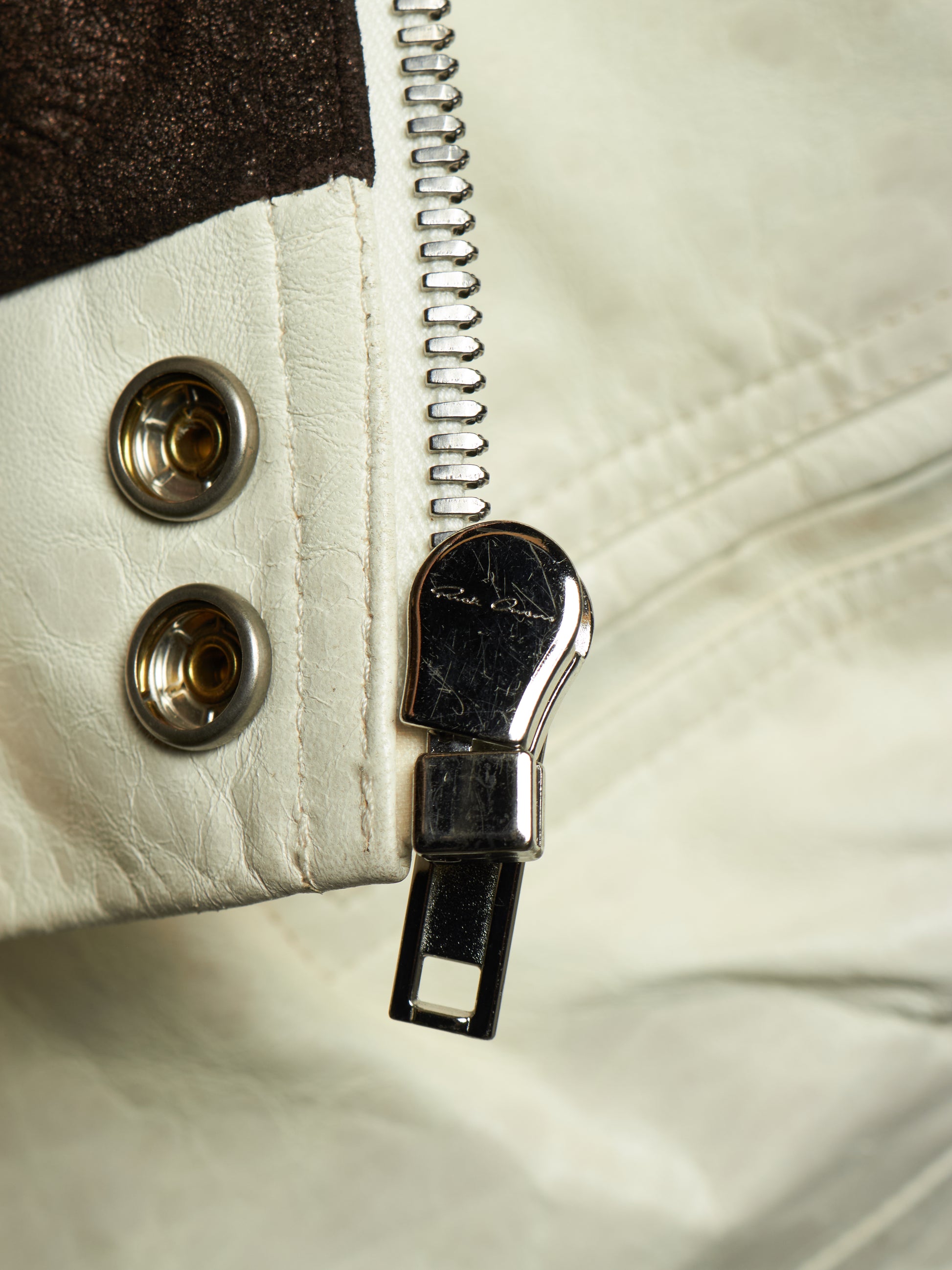 Close-up of a cream-colored leather jacket with a silver metal zipper, a dark brown textured panel, and two decorative silver snap buttons.