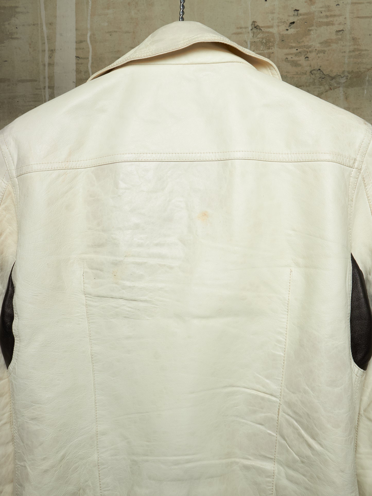 The back view of a cream-colored leather jacket hanging against a textured concrete wall, featuring dark brown leather sleeve accents.