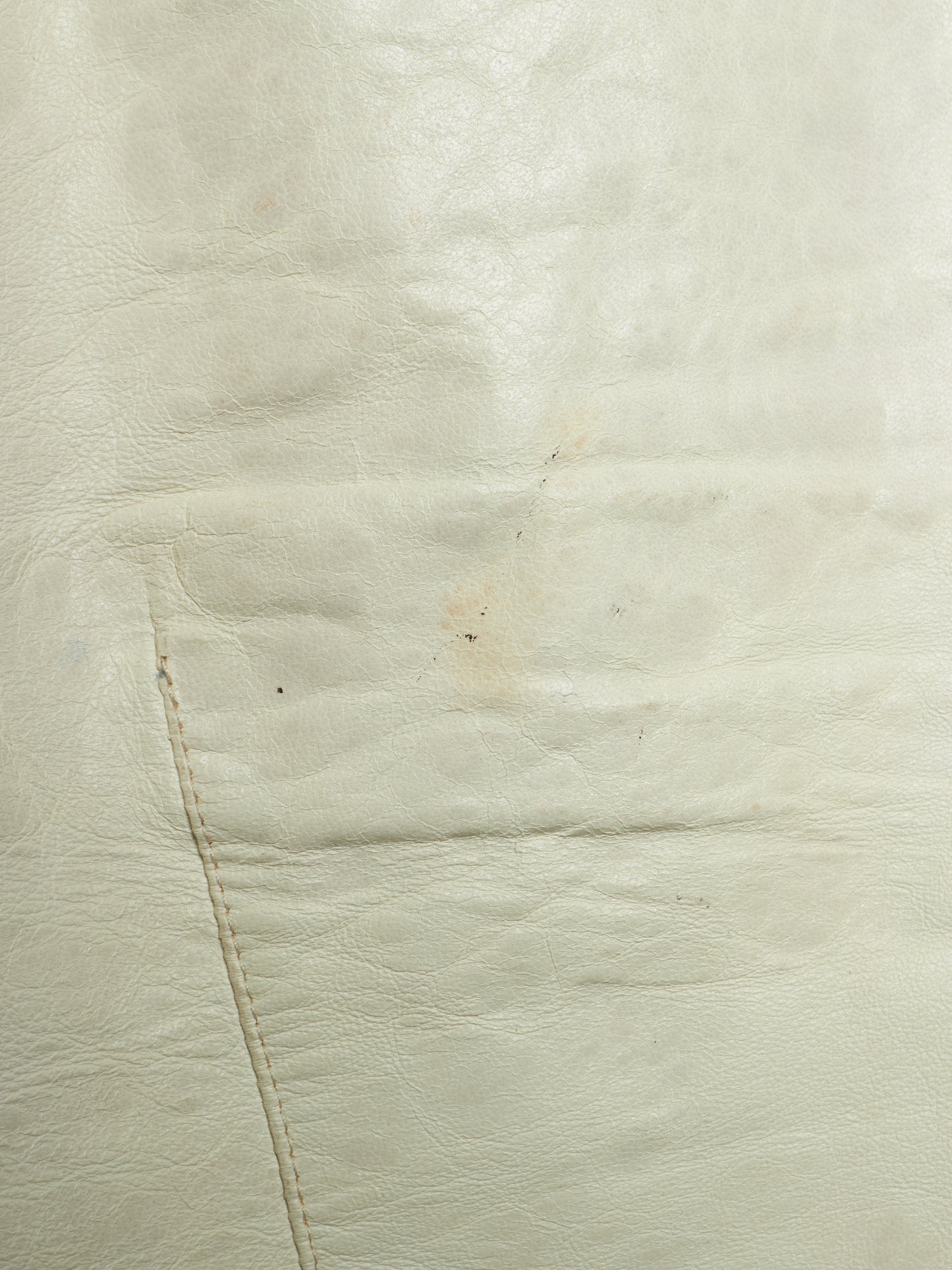 A close-up of a light cream or off-white leather surface with visible grain, stitching, and a small faint stain or discoloration.