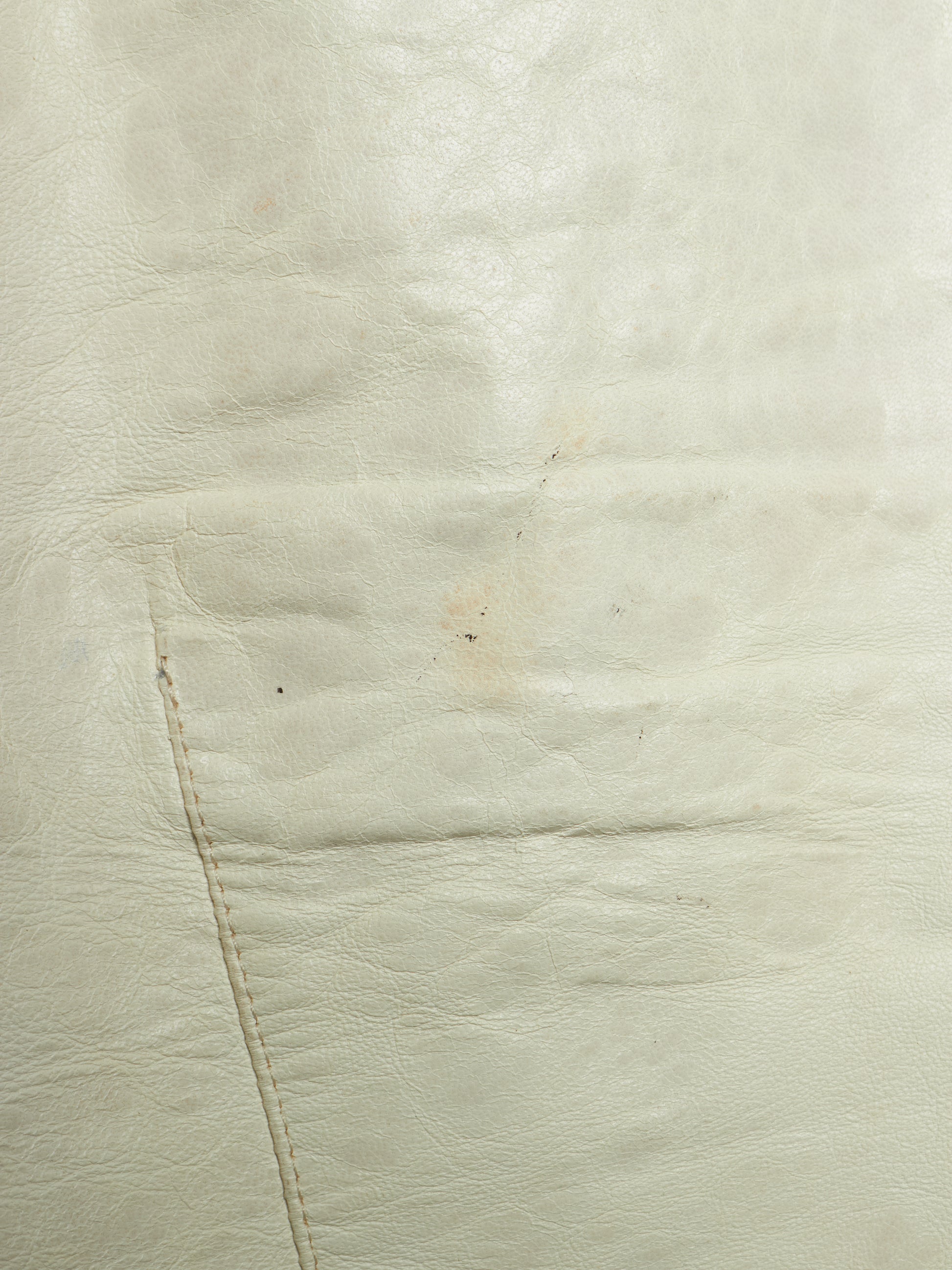 A close-up of a light cream or off-white leather surface with visible grain, stitching, and a small faint stain or discoloration.