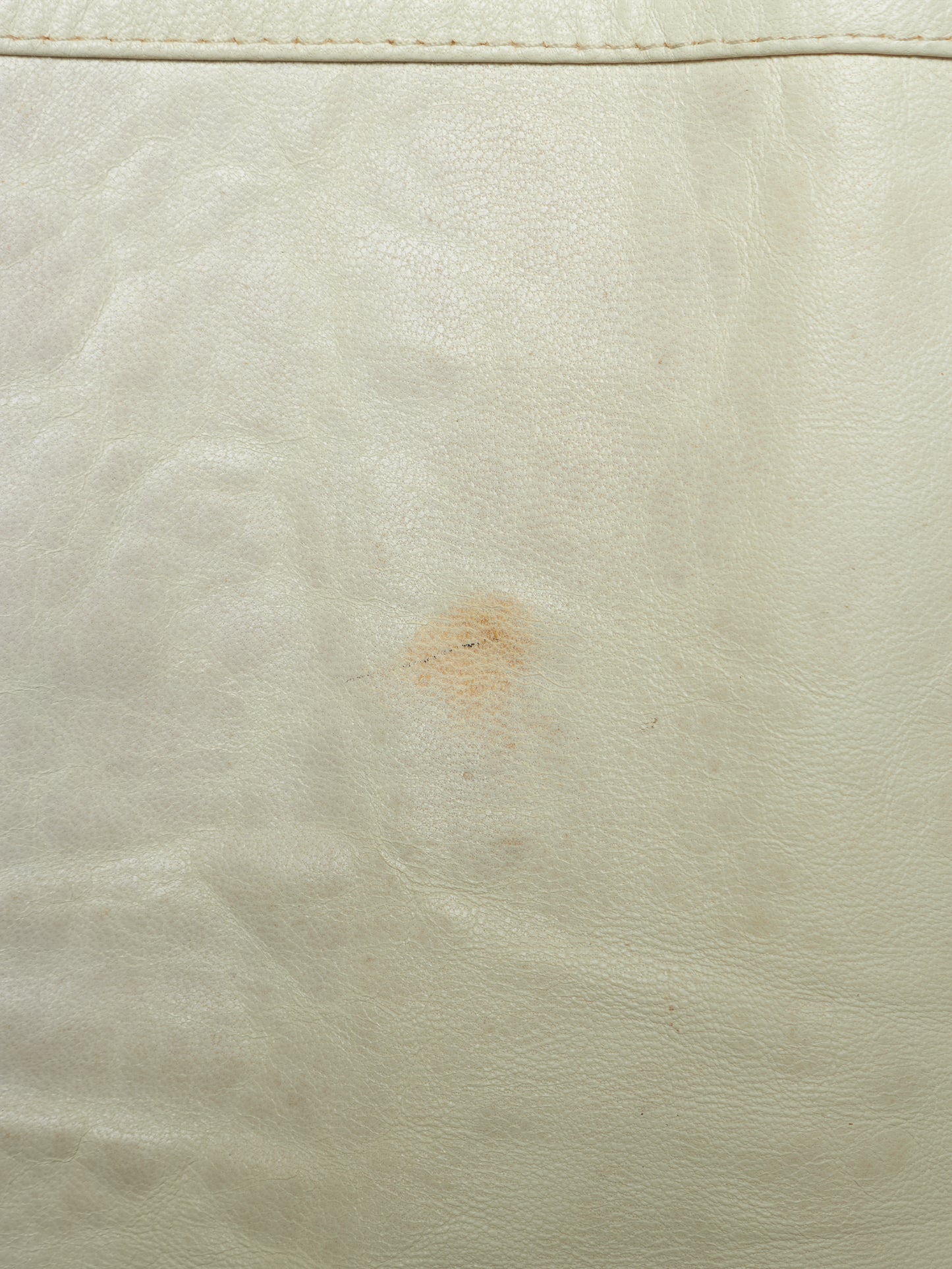 A close-up of a light cream or off-white leather surface with visible grain texture, a stitched seam near the top edge, and a small brownish stain in the center.