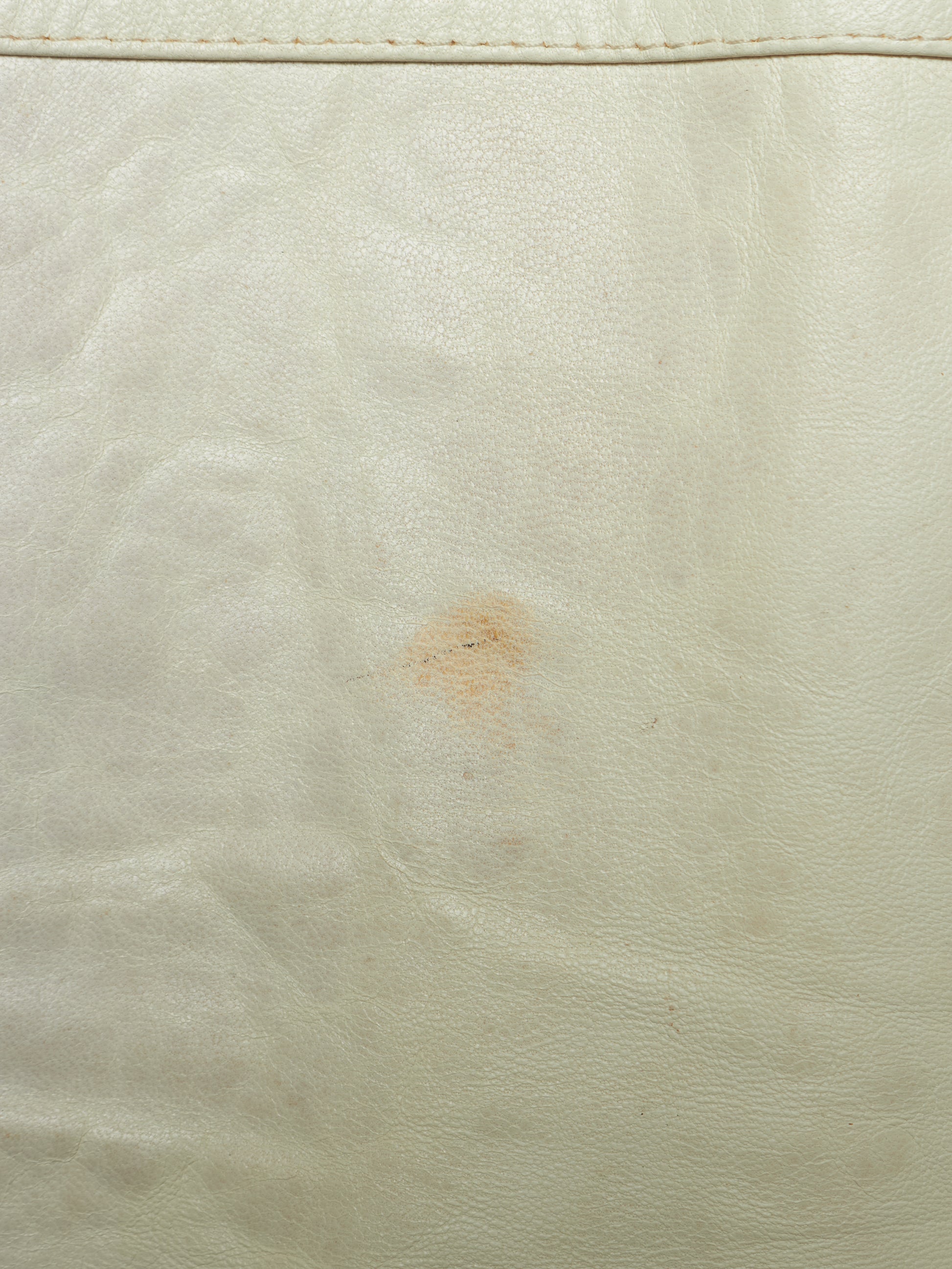 A close-up of a light cream or off-white leather surface with visible grain texture, a stitched seam near the top edge, and a small brownish stain in the center.
