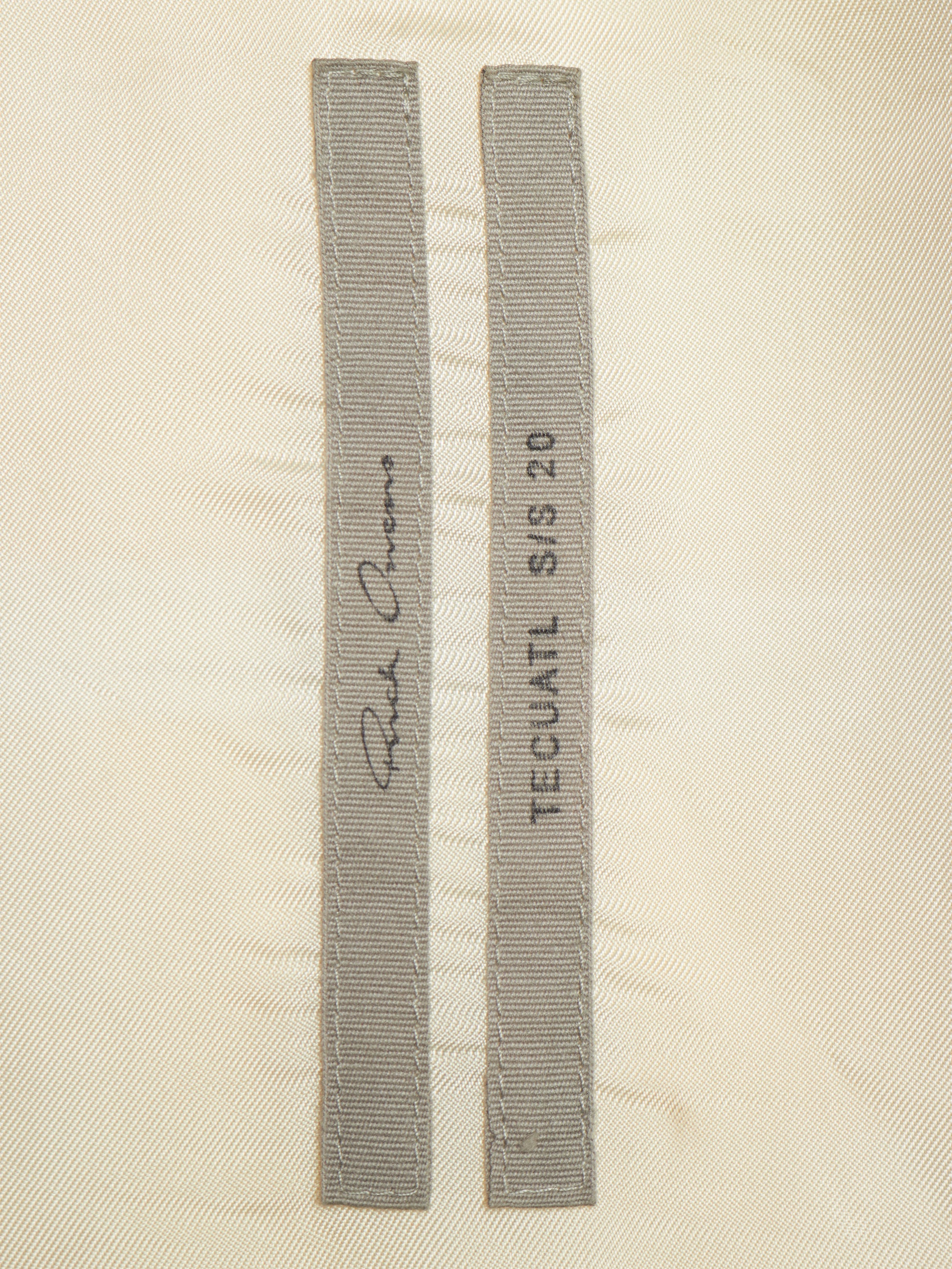Two narrow gray fabric strips with stitched edges laid flat on a light beige textured background. One strip has a cursive signature and the other reads 'TECUATL S/S 20'.