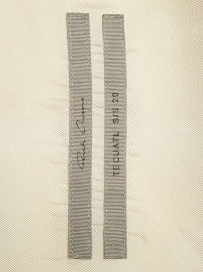 Two narrow gray fabric strips with stitched edges laid flat on a light beige textured background. One strip has a cursive signature and the other reads 'TECUATL S/S 20'.