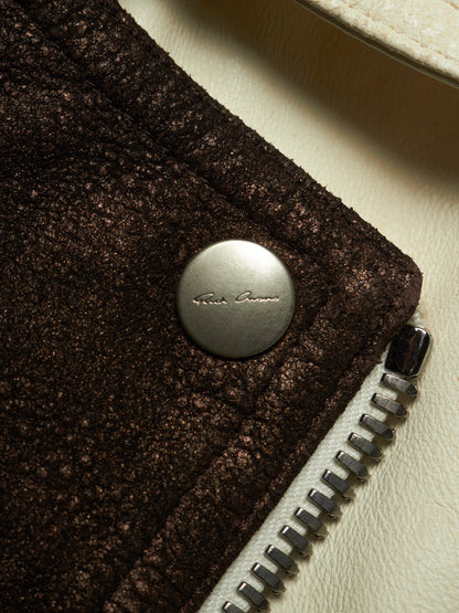 Close-up detail of a dark brown textured jacket featuring a silver-tone snap button with 'Rick Owens' branding and a silver zipper on a cream-colored leather background.