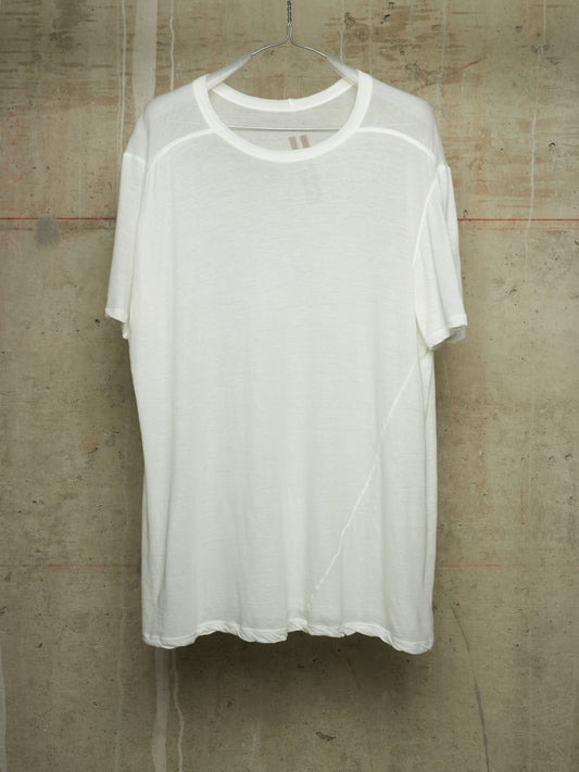 A white short-sleeve crewneck t-shirt with a sheer mesh overlay, hanging on a metal hanger against a textured concrete wall.