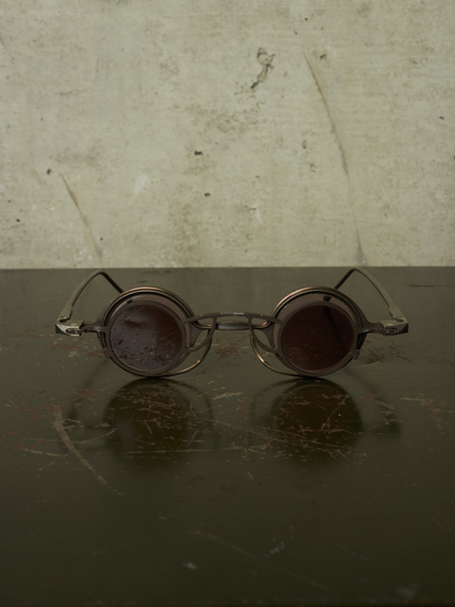 Vintage-style round metal eyeglasses with dark tinted lenses, resting on a dark reflective surface against a textured beige wall.