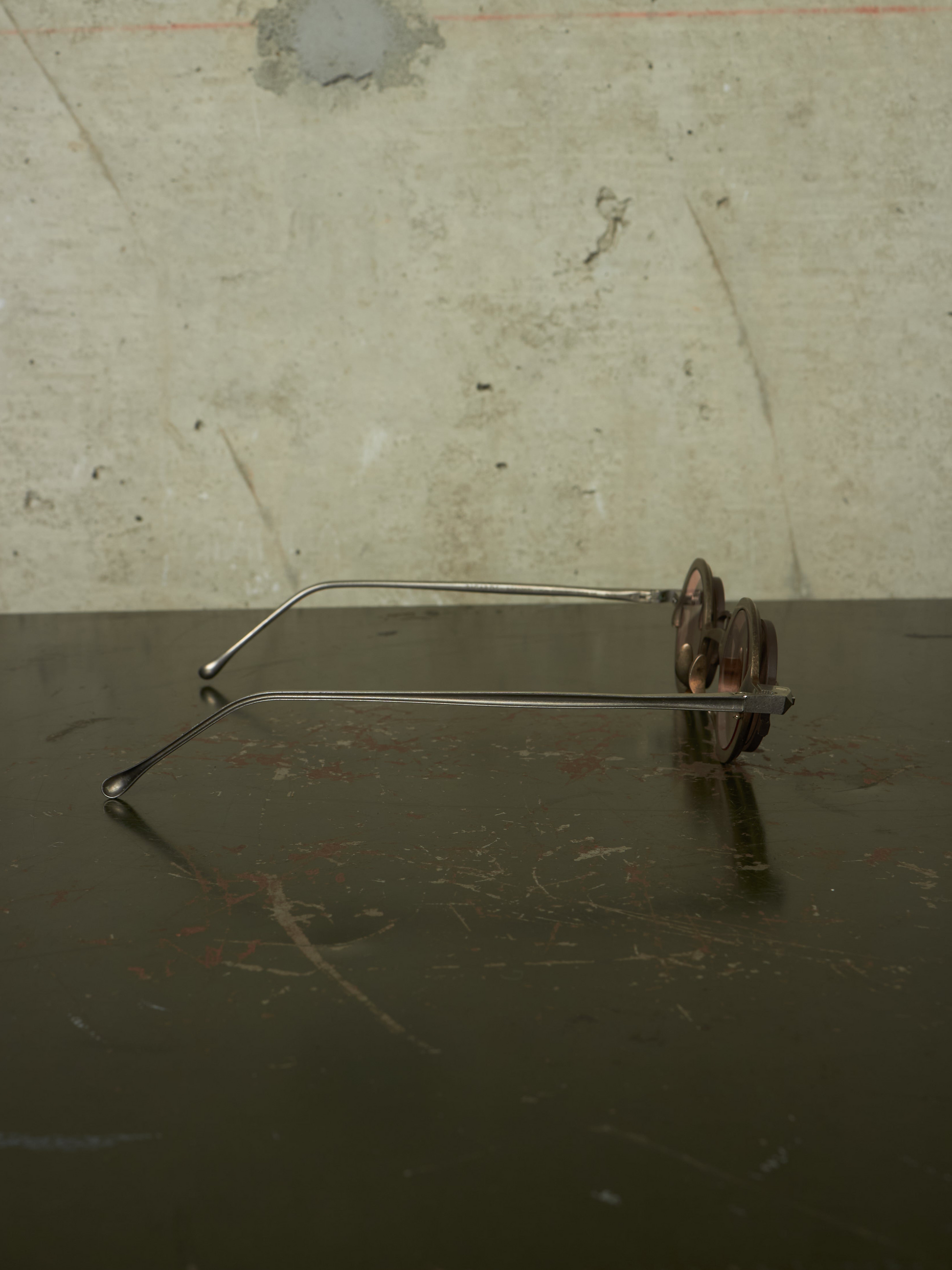 Vintage metal eyeglasses with a thin silver wire frame and dark brown decorative flower-shaped temples, resting on a dark reflective surface against a textured concrete wall.