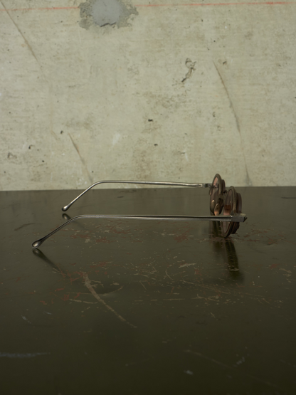 Vintage metal eyeglasses with a thin silver wire frame and dark brown decorative flower-shaped temples, resting on a dark reflective surface against a textured concrete wall.