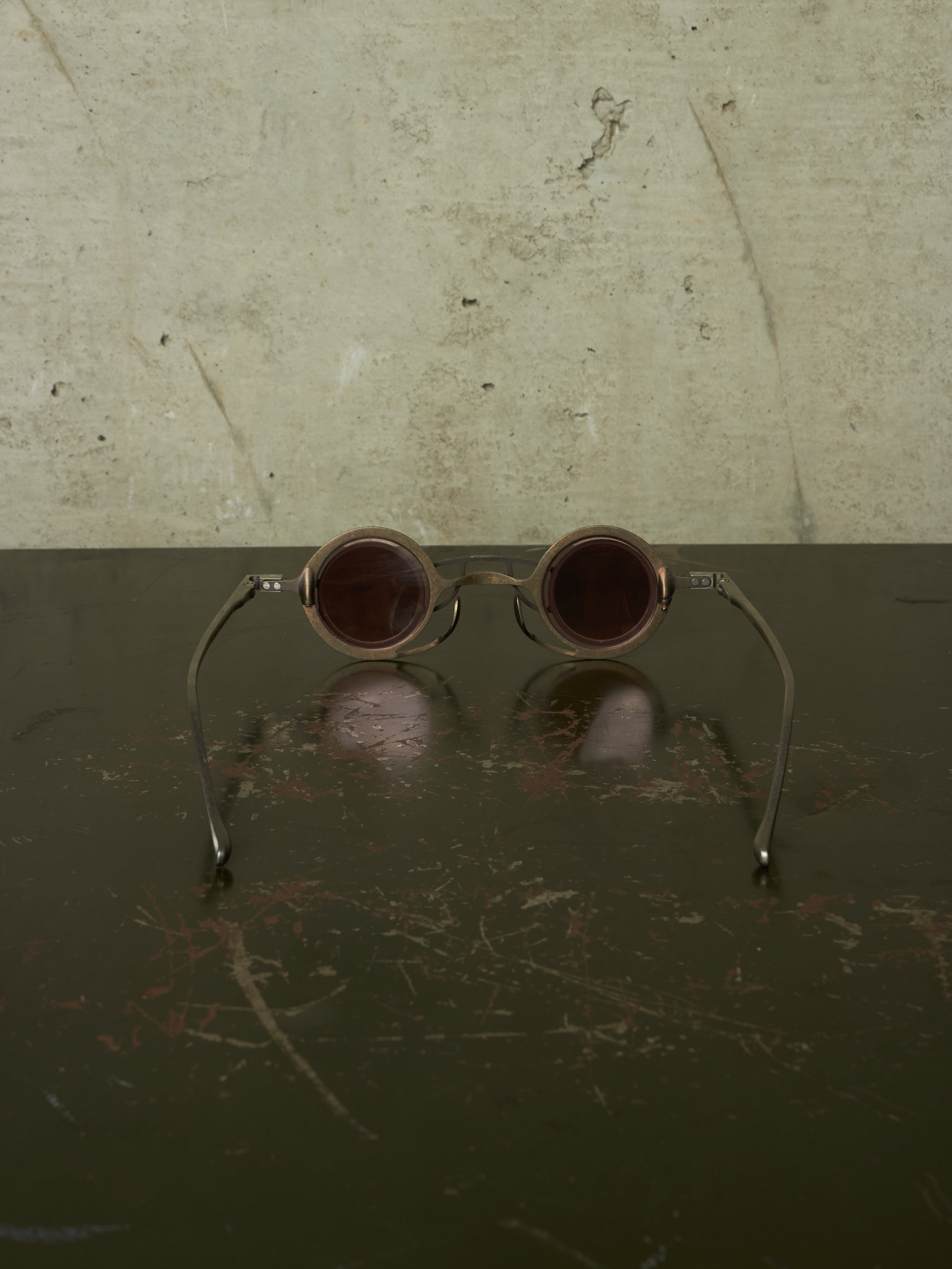 Round metal-framed sunglasses with dark brown tinted lenses resting on a dark reflective surface against a textured light beige wall.