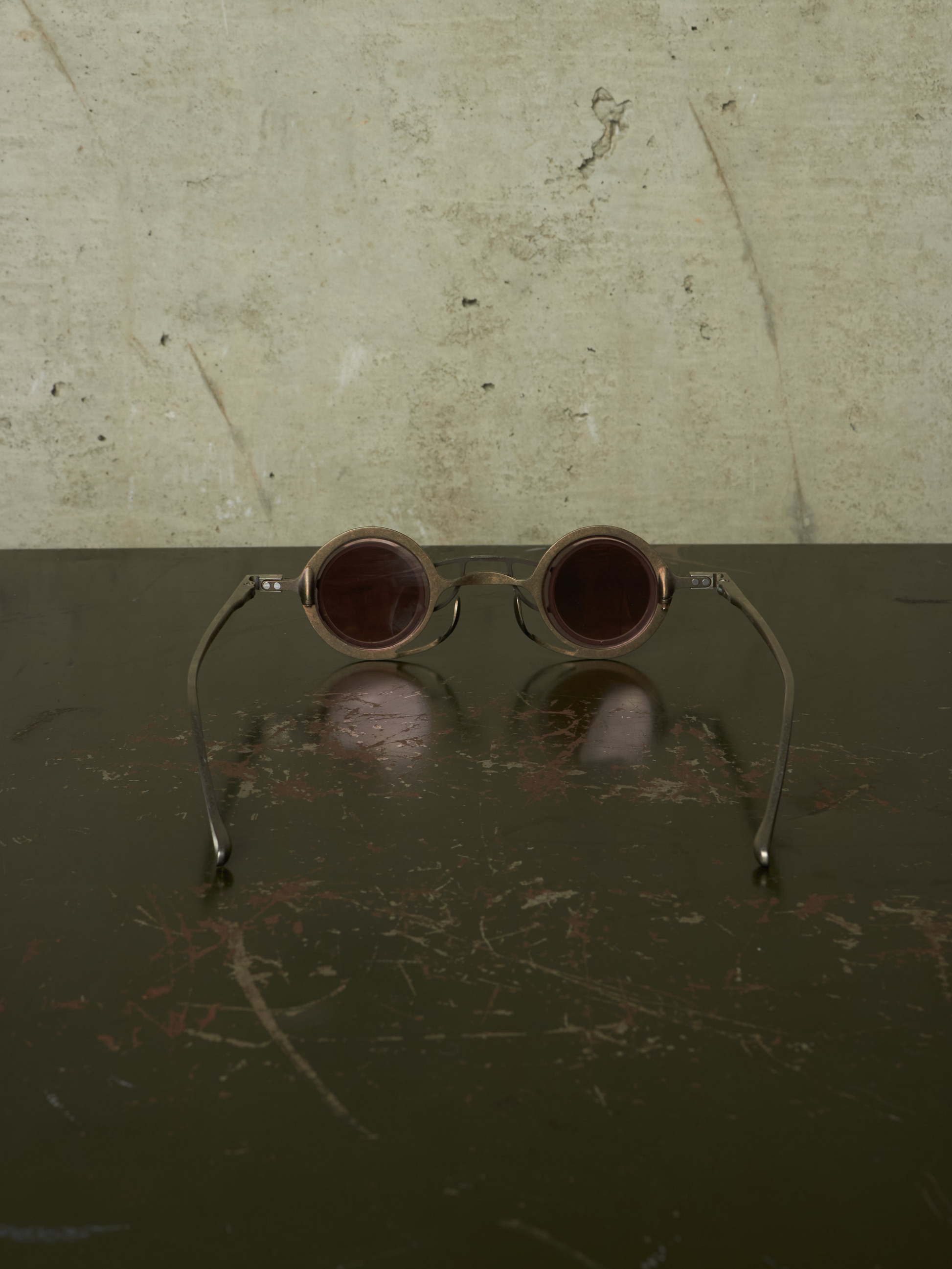 Round metal-framed sunglasses with dark brown tinted lenses resting on a dark reflective surface against a textured light beige wall.