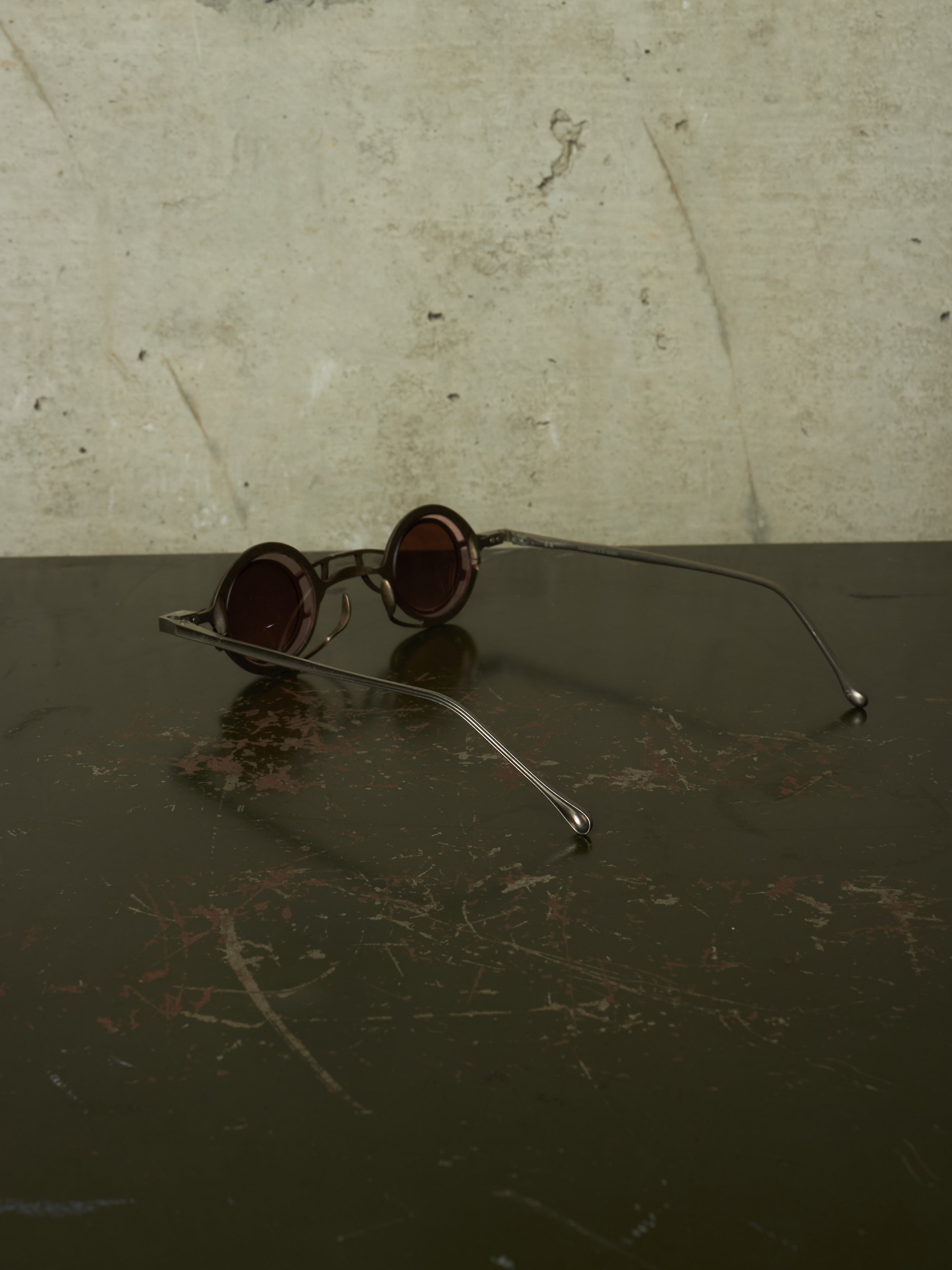 Round metal-frame sunglasses with dark tinted lenses resting on a dark reflective surface against a textured light beige wall.