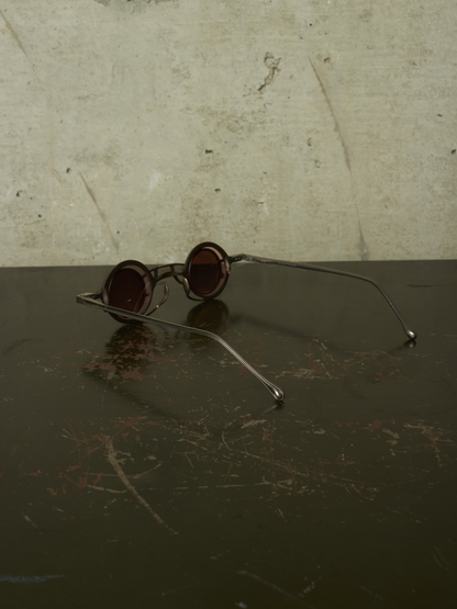 Round metal-frame sunglasses with dark tinted lenses resting on a dark reflective surface against a textured light beige wall.