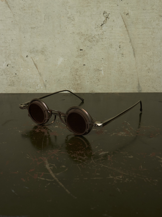 Round-frame metal eyeglasses with dark tinted lenses resting on a dark reflective surface against a textured light wall.