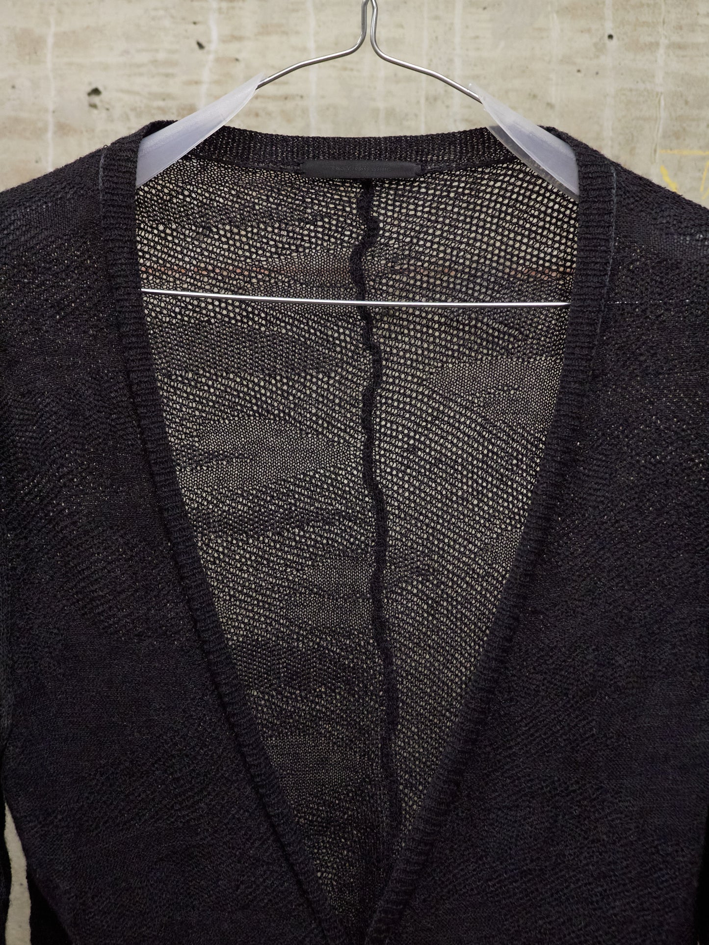 Black cardigan with a visible tear on a hanger against a beige tiled wall.