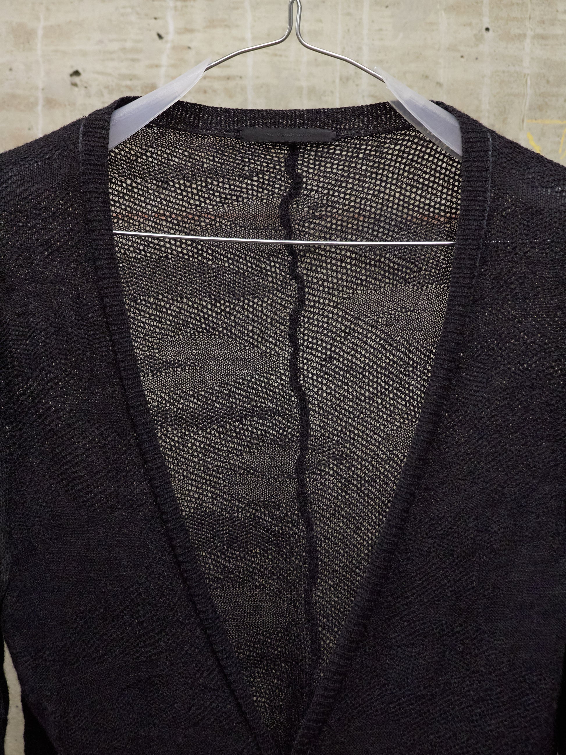 Black cardigan with a visible tear on a hanger against a beige tiled wall.
