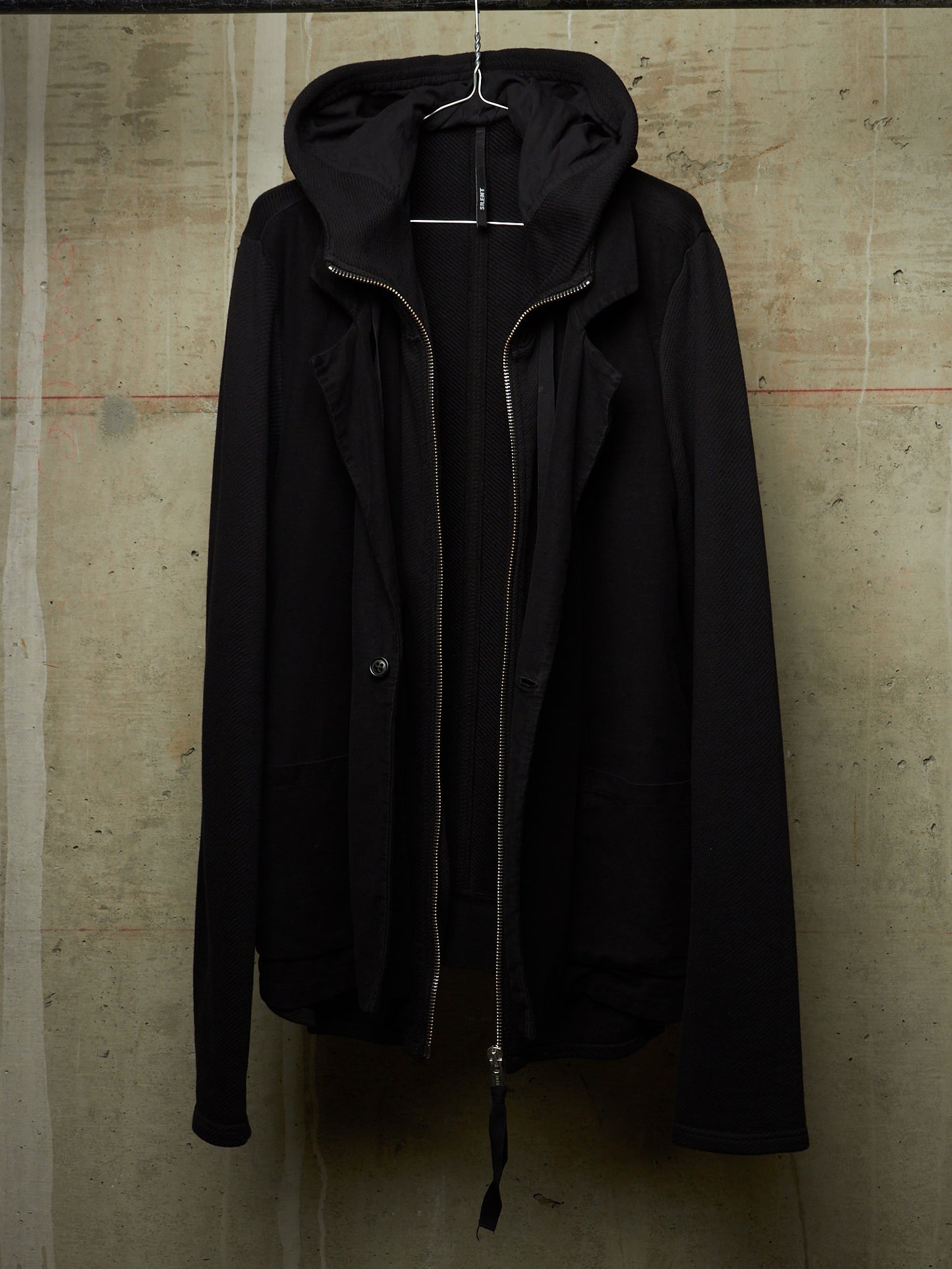 A black hooded jacket with a front zipper and snap button closure, hanging on a wire hanger against a textured concrete wall.