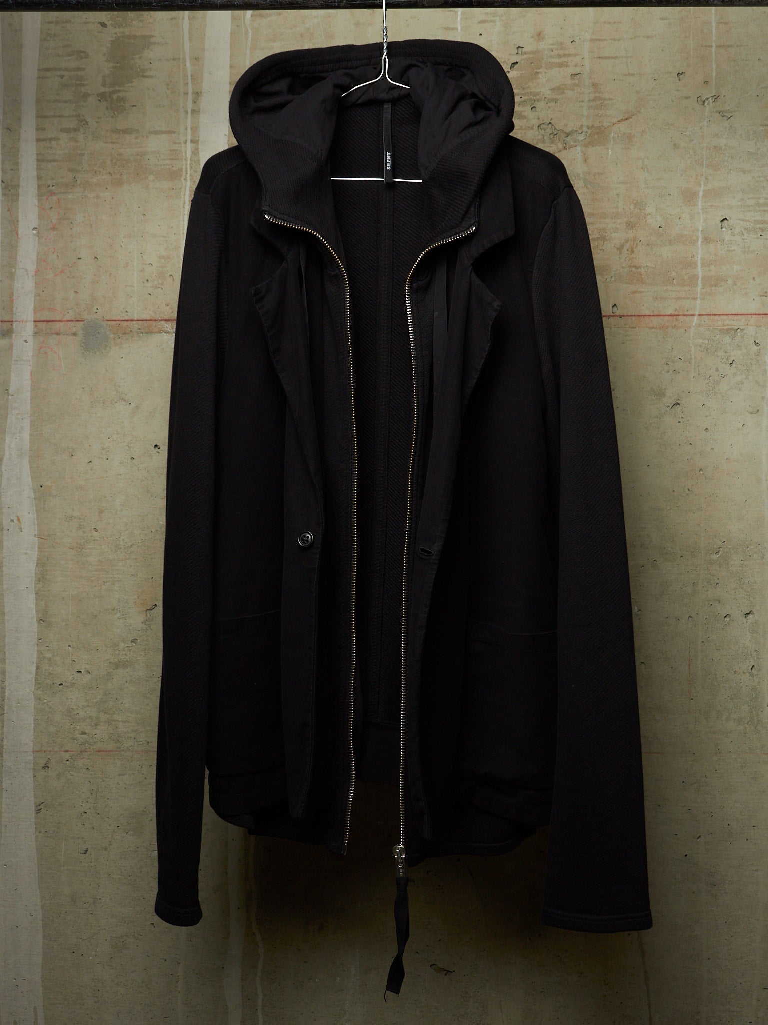 A black hooded jacket with a front zipper and snap button closure, hanging on a wire hanger against a textured concrete wall.