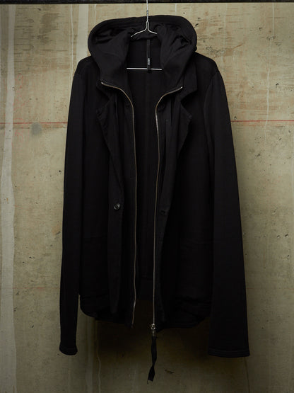 A black hooded jacket with a front zipper and snap button closure, hanging on a wire hanger against a textured concrete wall.