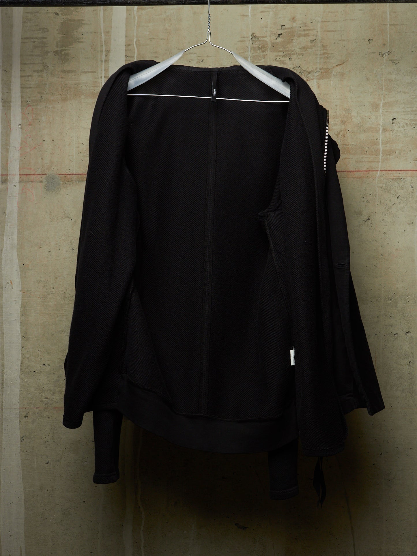A black zip-up jacket hanging on a white hanger against a textured concrete wall.