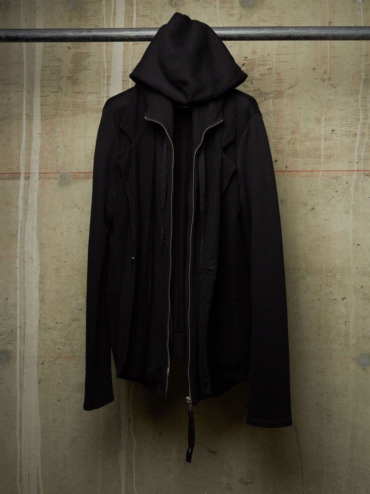 A black hooded jacket with a front zipper, hanging from a metal bar against a textured concrete wall.