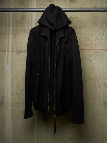 A black hooded jacket with a front zipper, hanging from a metal bar against a textured concrete wall.