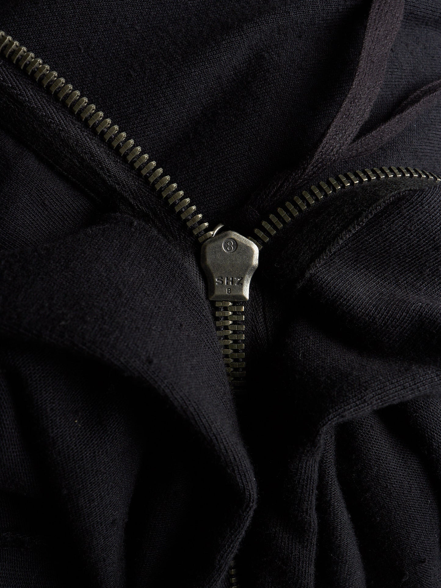 Close-up of a black zippered garment with a dark metal zipper pull and zipper teeth, showing the fabric texture and construction details.