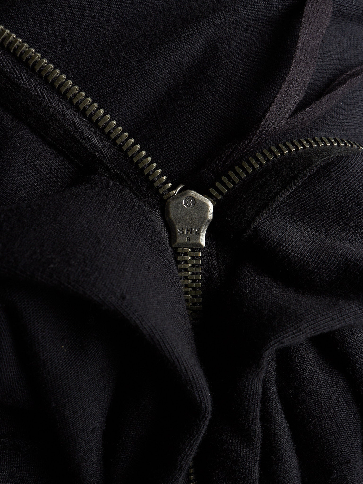 Close-up of a black zippered garment with a dark metal zipper pull and zipper teeth, showing the fabric texture and construction details.