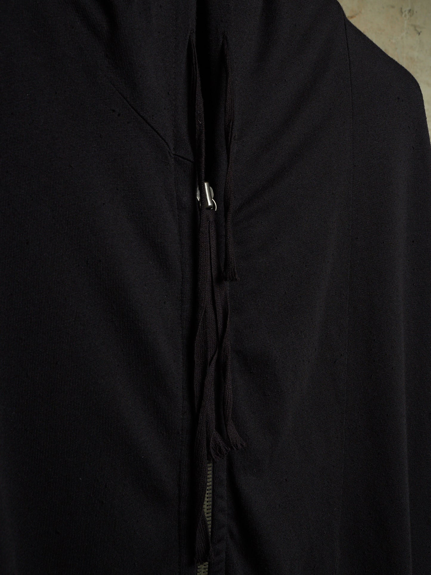 Close-up of a black garment with a front zipper and drawstring detail, showing the fabric texture and a small silver zipper pull.