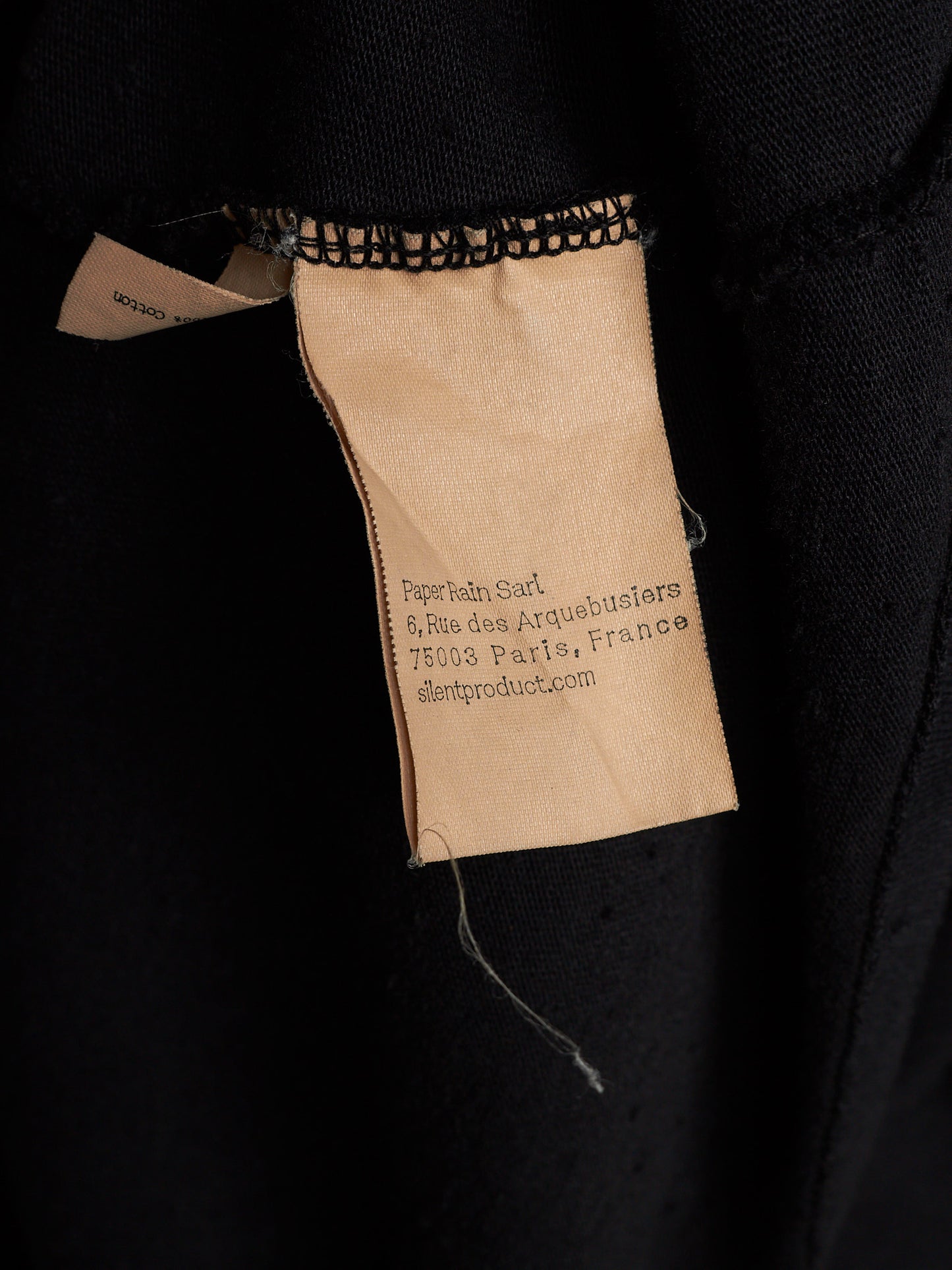 Close-up of a black garment showing a sewn-on paper brand label that reads 'Perthain Seld. 6,Rue des Arquebusiers 75003 Paris, France' and 'sleptomproduct.