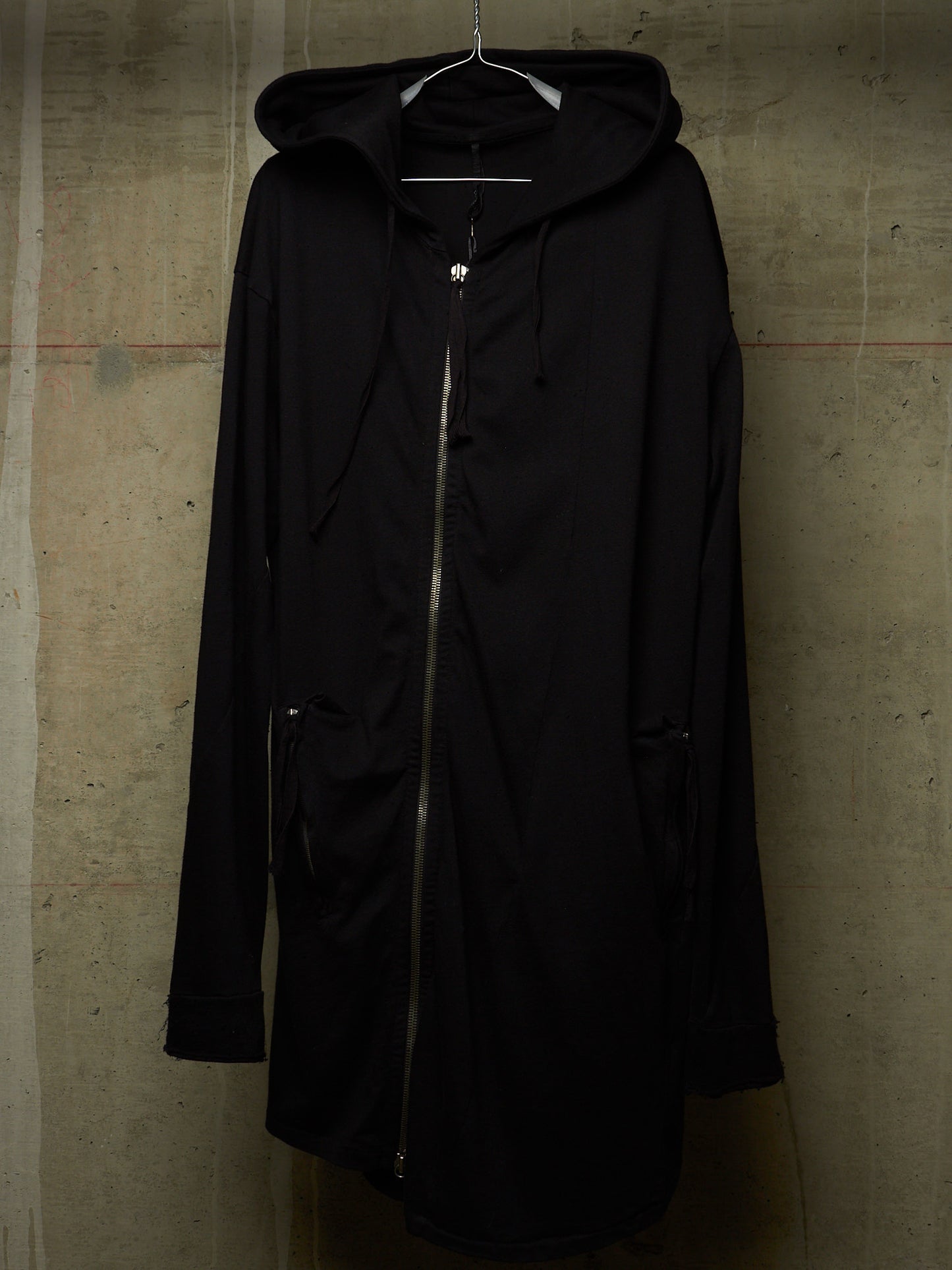 A black hooded zip-up coat with drawstrings and side pockets, hanging on a metal hanger against a textured concrete wall.