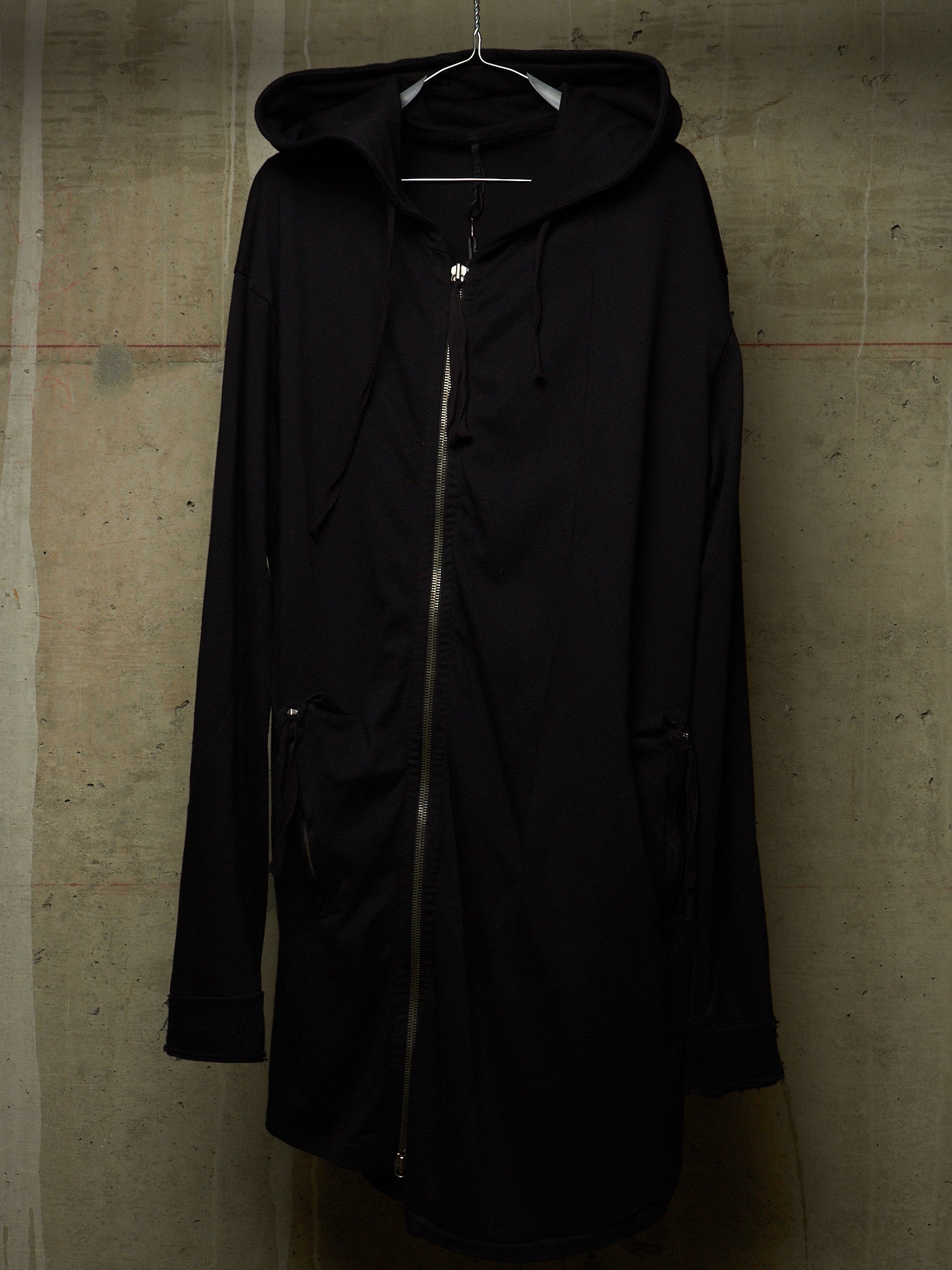 A black hooded zip-up coat with drawstrings and side pockets, hanging on a metal hanger against a textured concrete wall.