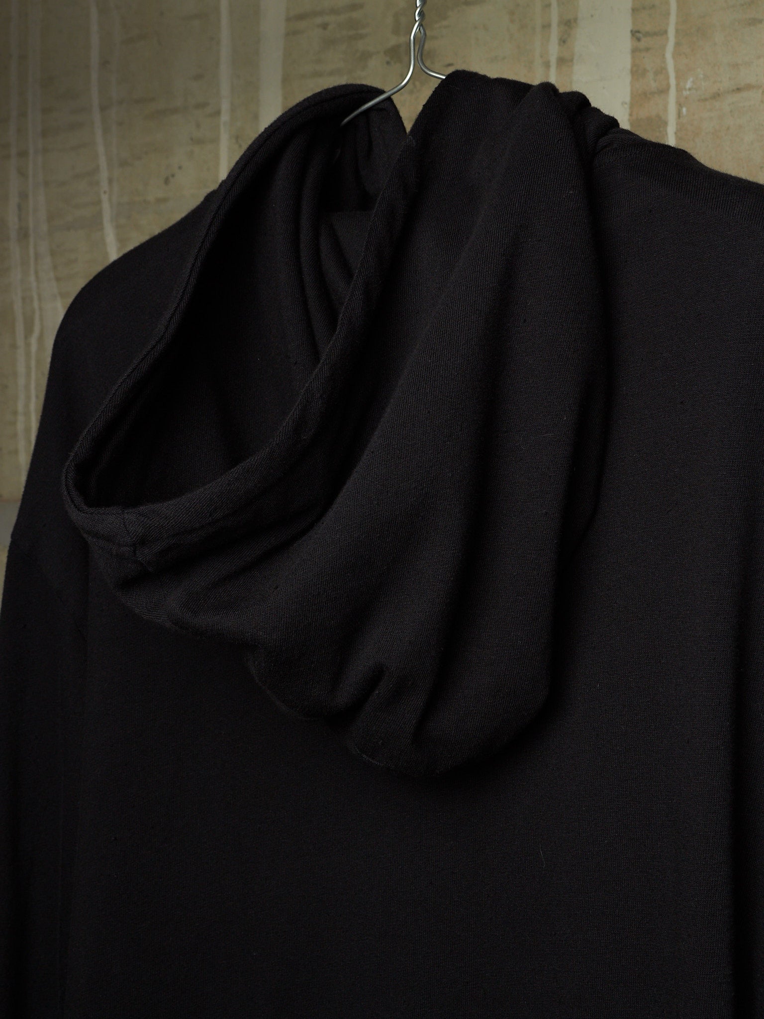 A black draped cowl-neck top or hoodie hanging on a metal hanger against a textured beige wall.