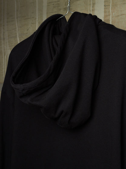 A black draped cowl-neck top or hoodie hanging on a metal hanger against a textured beige wall.