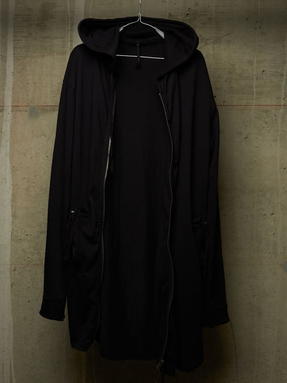 A black hooded jacket with a front zipper, hanging on a metal hanger against a textured concrete wall.
