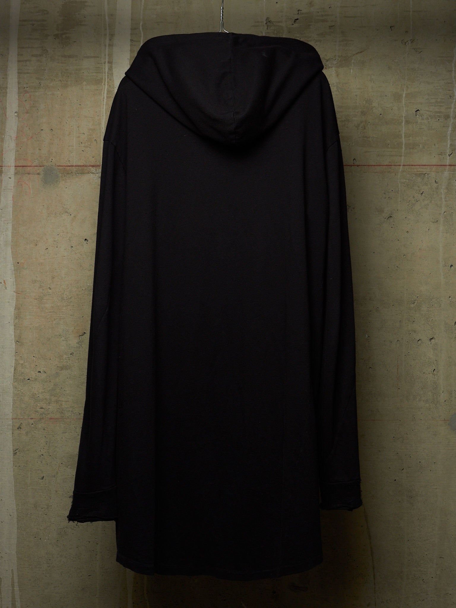 A black hooded long-sleeve garment, shown from the back, hanging against a textured concrete wall.
