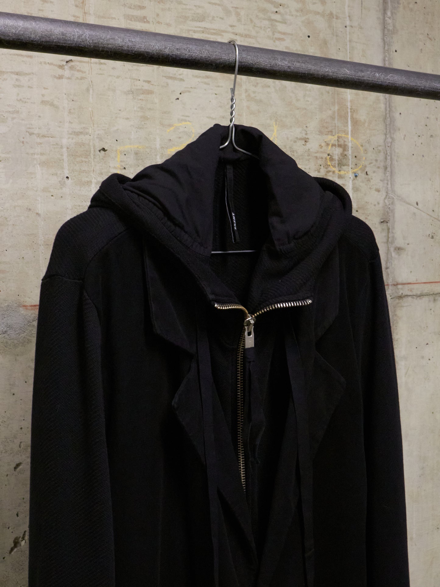 Black jacket with a gold zipper on a hanger against a concrete wall.