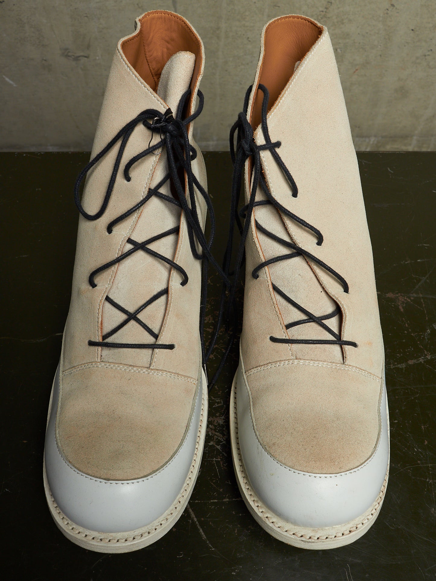 A pair of beige suede lace-up ankle boots with white leather toe caps and black laces, displayed on a dark surface against a concrete wall.