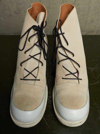 A pair of beige suede lace-up ankle boots with white leather toe caps and black laces, displayed on a dark surface against a concrete wall.