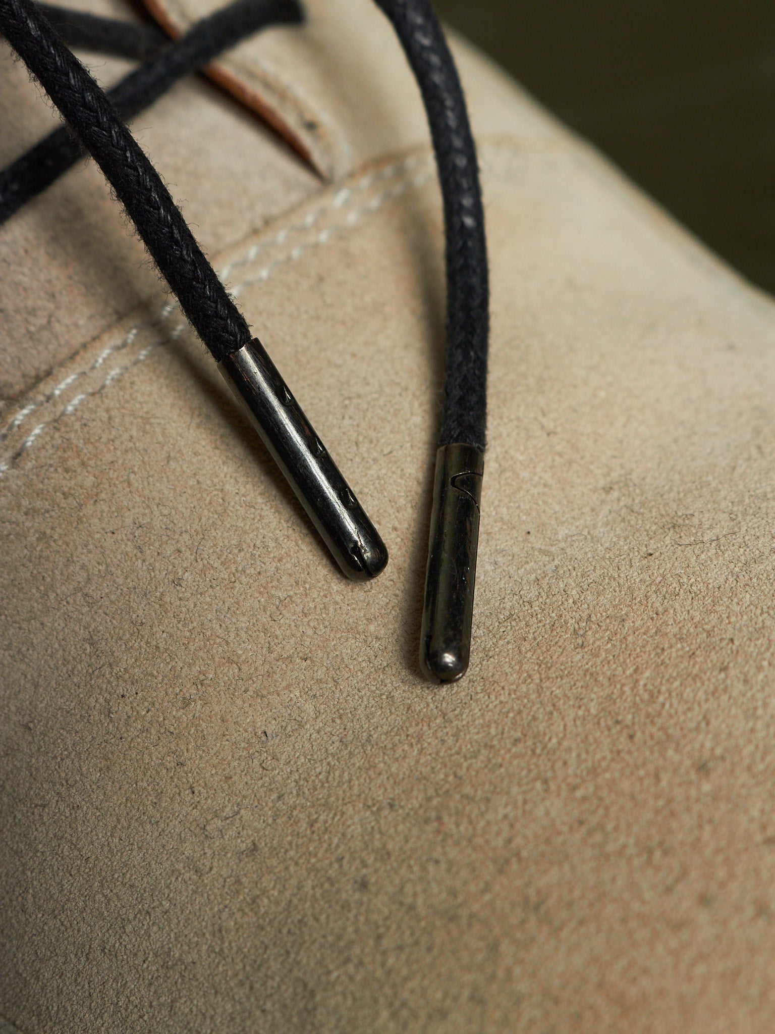 Close-up of black braided laces with dark metal aglets attached to a beige suede or leather shoe surface.
