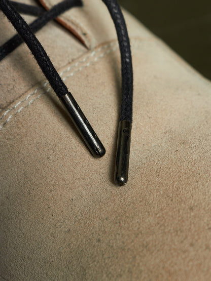 Close-up of black braided laces with dark metal aglets attached to a beige suede or leather shoe surface.