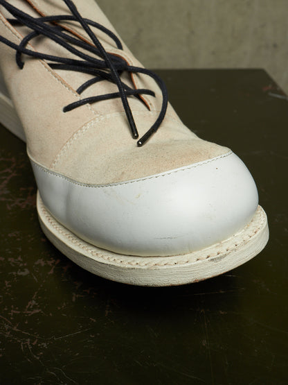 A close-up of a low-top lace-up boot with a cream suede upper, white leather toe cap, and off-white sole, resting on a dark surface.