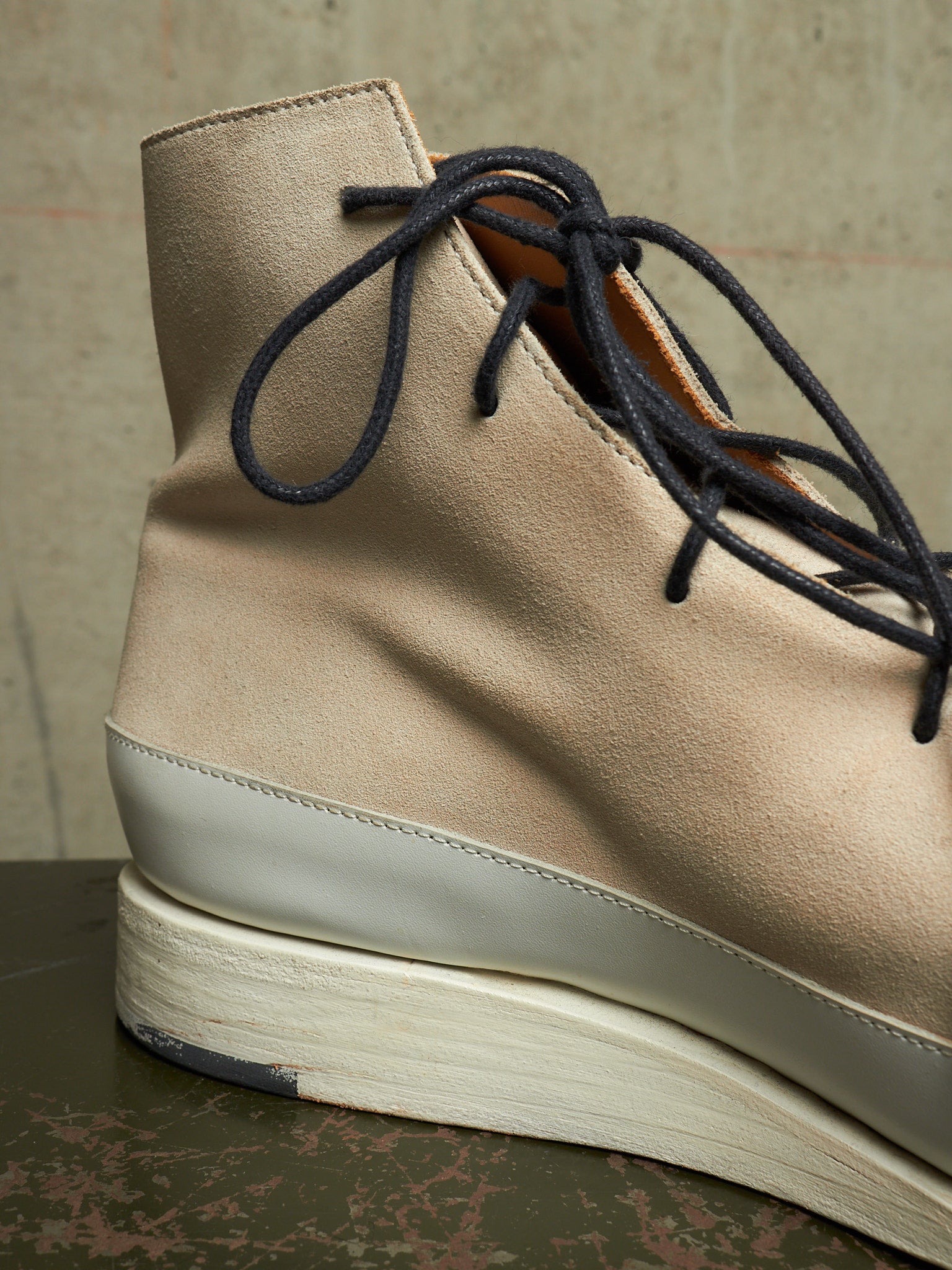 A close-up of a beige suede ankle boot with dark navy laces, a thick off-white midsole, and a textured dark green surface beneath it.