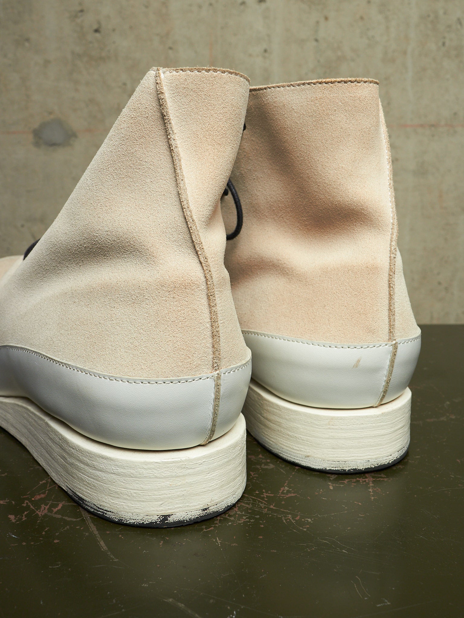 A pair of beige suede ankle boots with white rubber soles, shown from the back on a dark tabletop against a textured concrete wall.