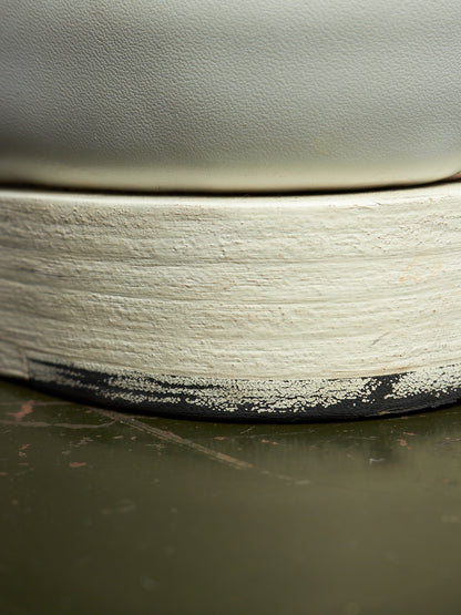 Close-up of a ceramic vessel showing a thick, textured off-white painted rim with a black unglazed edge, resting on a dark olive-green surface.