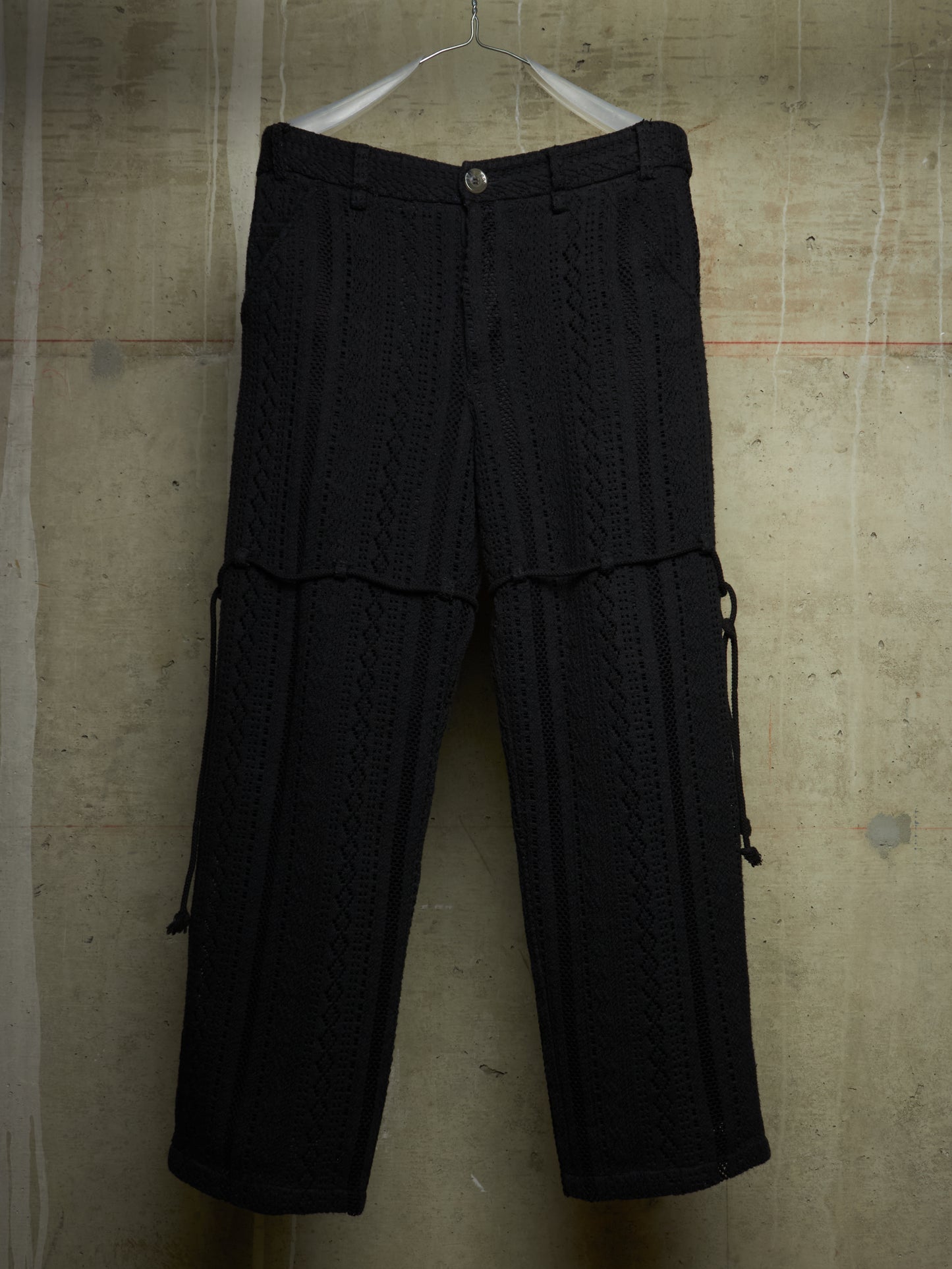 Black textured knit cargo pants with a button waist and side drawstring details, hanging on a white hanger against a concrete wall.