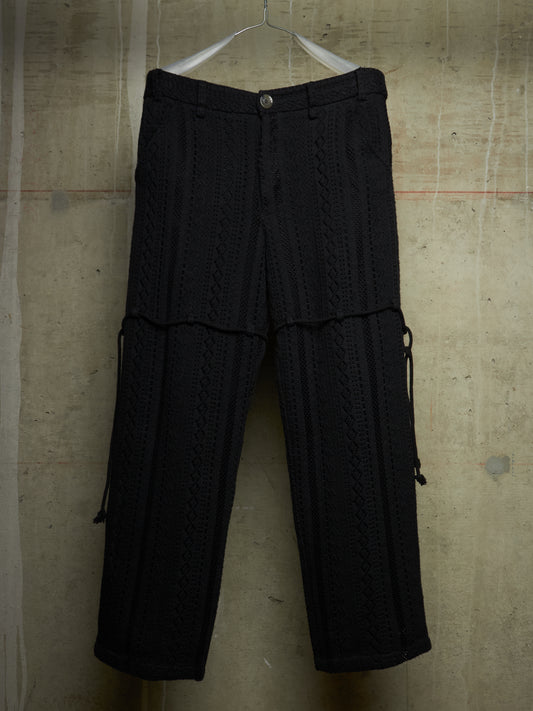 Black textured knit cargo pants with a button waist and side drawstring details, hanging on a white hanger against a concrete wall.