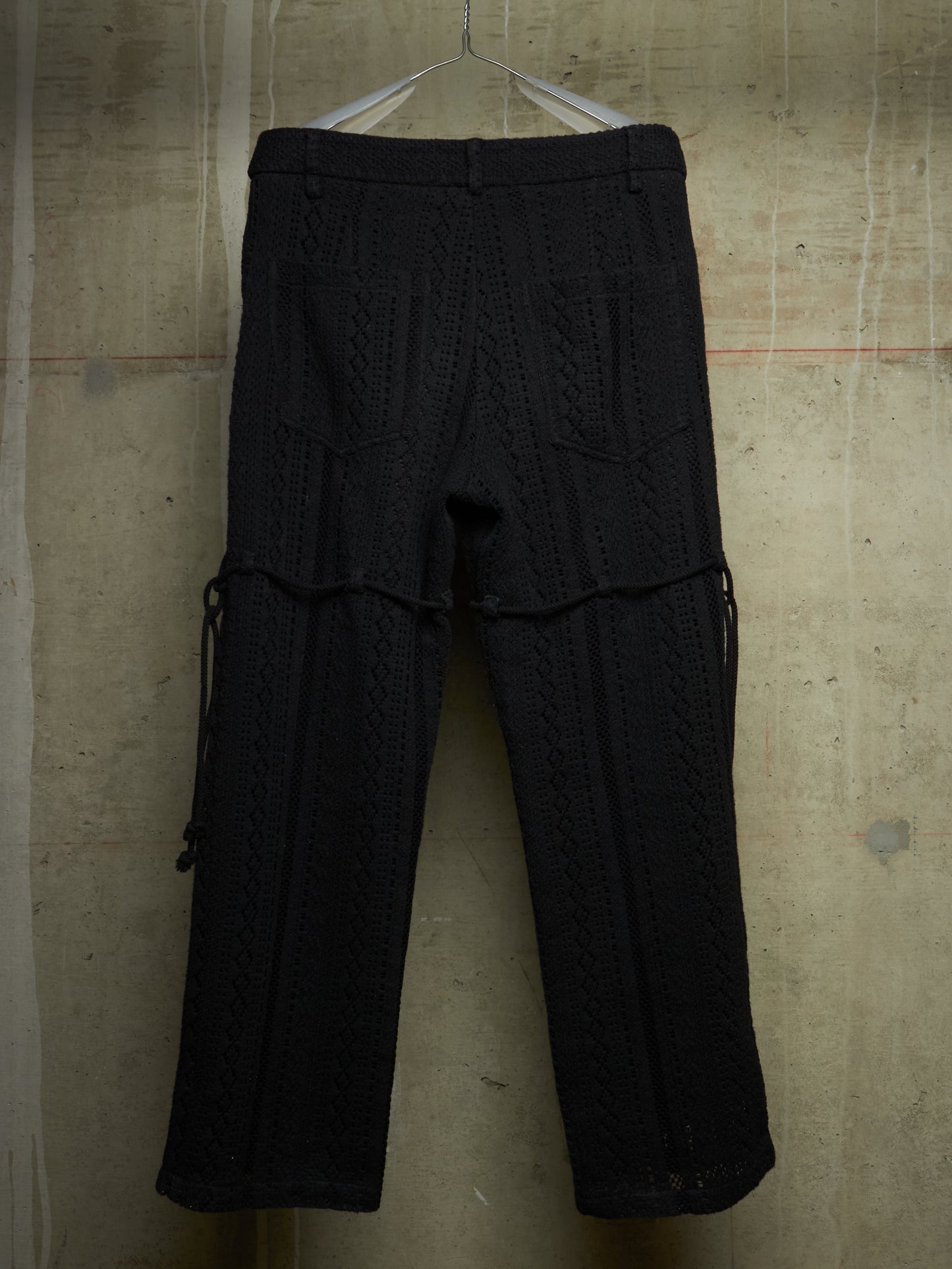 Black textured knit pants with side drawstrings, hanging on a white hanger against a concrete wall.