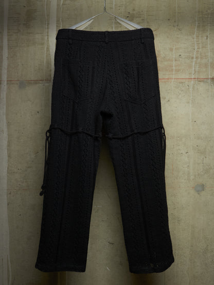 Black textured knit pants with side drawstrings, hanging on a white hanger against a concrete wall.