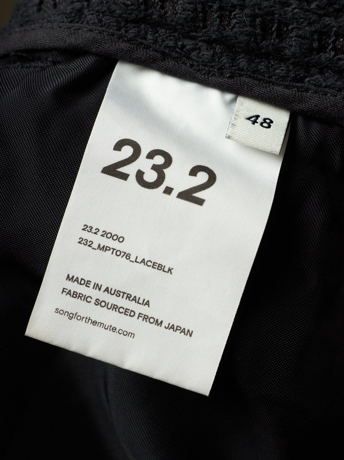Close-up of a black garment interior with a white size tag reading 23.2 and 48, plus text noting made in Australia and fabric sourced from Japan.
